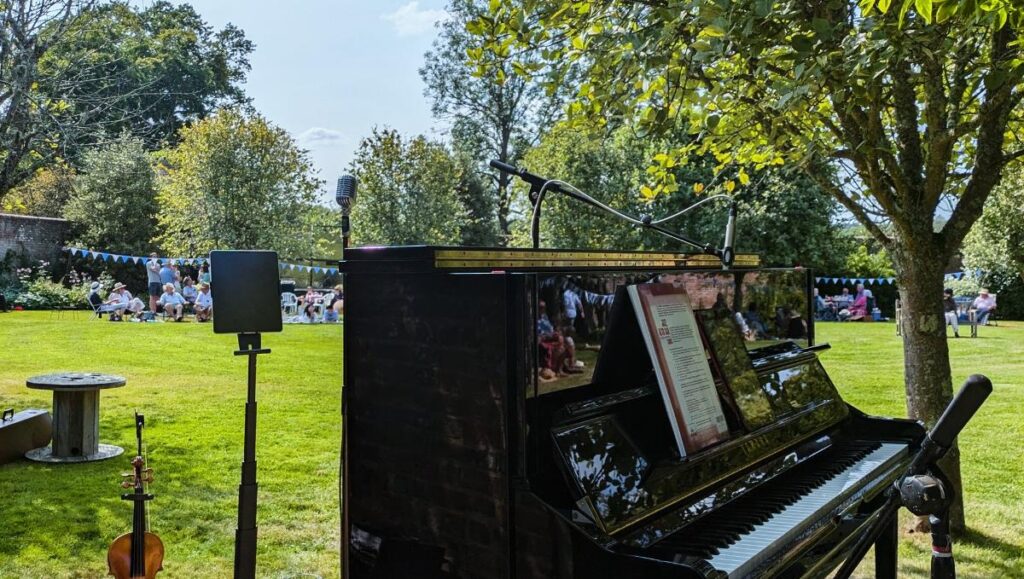 View from the stage in the walled garden. A violin is in the foreground, to the left. A music stand centre stage and a mic-ed up acoustic Yamaha to the right. In the background is the walled garden decorated with bunting.