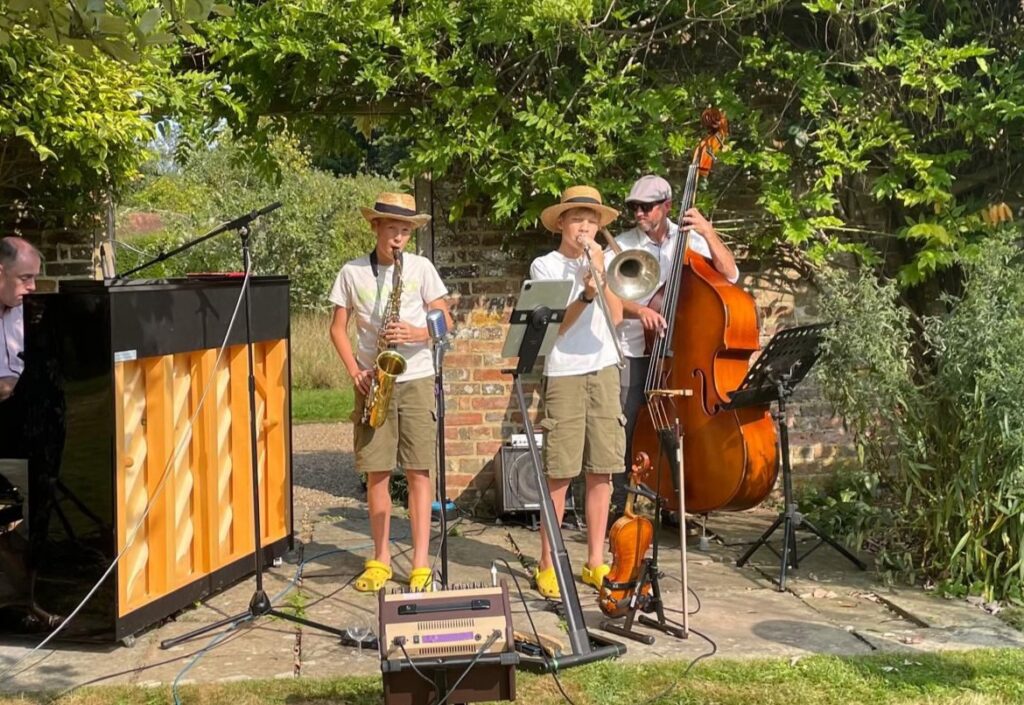 Theo and Reuben Nichols performing Watermelon Man on saxophone and trombone accompanied by Daivd Hall (left) on an acoustic Yamaha and James Sunney on double bass (right). Behind them is wall of the walled garden, covered in wisteria.