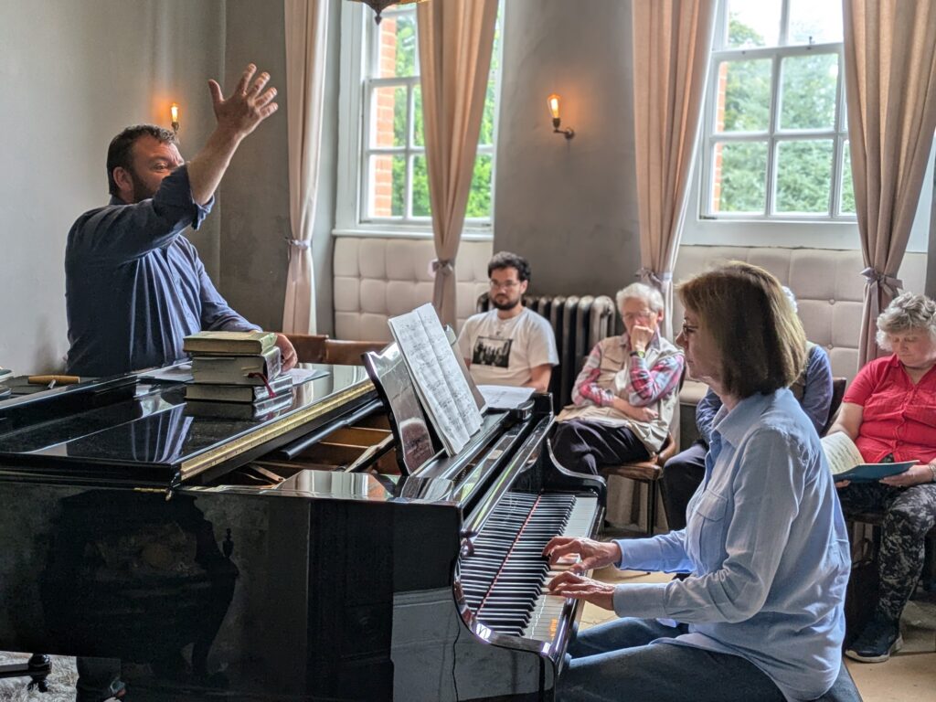 Pianist playing a Steinway model B in a performance class. The tutor stands behind the piano to the left and is demonstrating energetically with his arm to encourage her. Other students watch on in the background.