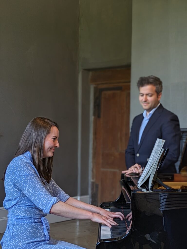 A young woman plays a Steinway model B in a blue dress. She smiles as she plays. A teachers listens on in the background, also smiling. He wears a navy suit and light blue shirt.