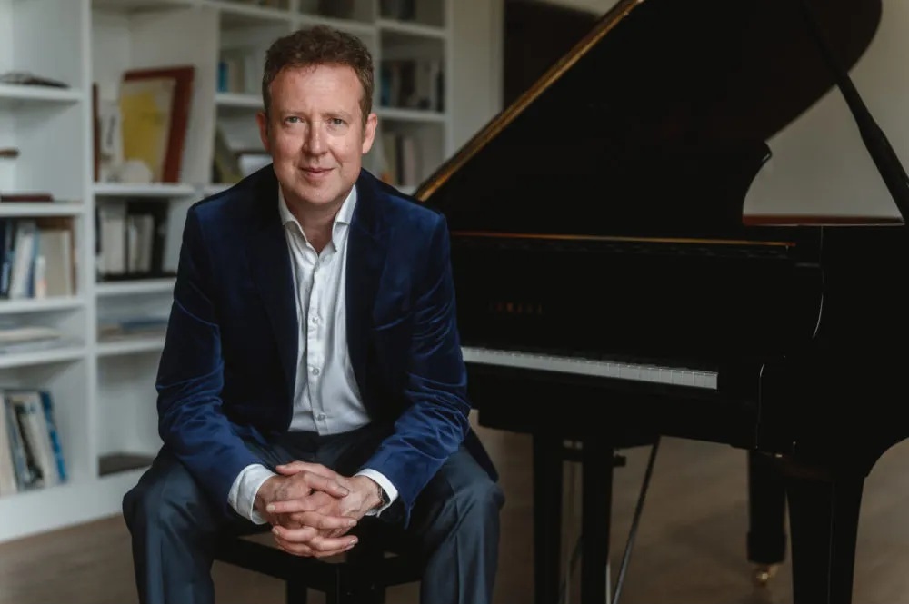 Sam Haywood sits on piano stool smiling at the camera. He rests his elbows on his knees and interlaces his fingers. Behind him is a grand piano.