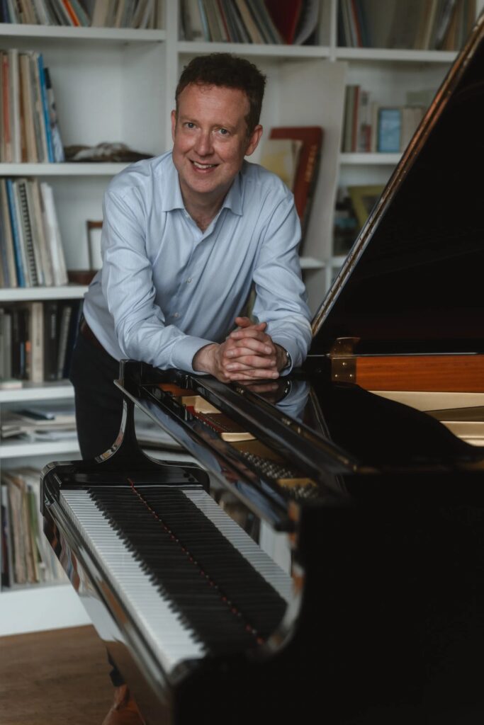 Sam Haywood leans forward against a grand piano smiling at the camera. He rests his elbows on the piano, his fingers interlaced. He wears a shirt and behind him is a white bookshelf containing sheet music.