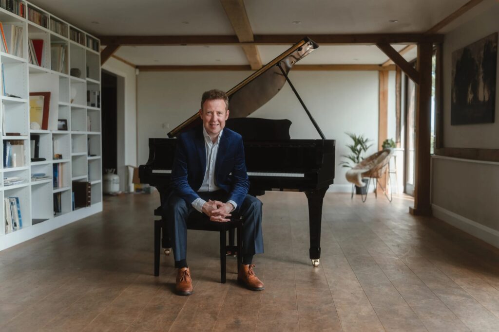 Sam Haywood sits in a large room on piano stool smiling at the camera. He rests his elbows on his knees and interlaces his fingers. Behind him is a grand piano. The floor is wooden and the room is supported by wall and ceiling wooden beams. There is a large white bookshelf to the left.